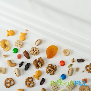 May include: A variety of snack foods on a white surface, including pretzels, goldfish crackers, cereal, nuts, dried fruit, and candy.