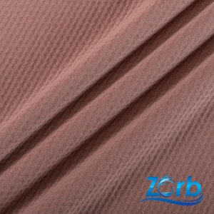 Puede incluir: Primer plano de una tela texturizada de color malva. La tela tiene un patr&oacute;n tejido sutil. La tela est&aacute; doblada por la mitad, creando una l&iacute;nea diagonal a trav&eacute;s de la imagen. La palabra "Zorb" es visible en la esquina inferior derecha de la imagen.