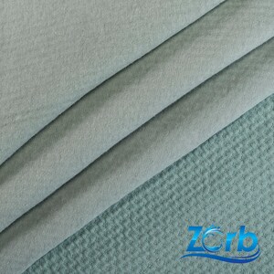 May include: A close-up of a light blue fabric with a waffle texture. The fabric is folded in thirds, with the top layer folded over the bottom two layers. The fabric is soft and smooth to the touch.