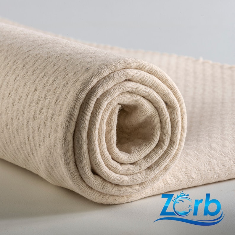 Puede incluir: Un primer plano de un rollo de tela de color natural con una superficie texturizada. La tela est&aacute; enrollada apretadamente y los bordes son visibles. La tela est&aacute; etiquetada con la palabra "Zorb".