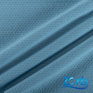 May include: A close-up of a blue waffle weave fabric. The fabric has a textured surface and a subtle sheen. The fabric is folded in half, creating a diagonal line across the image. The word "Zorb" is visible in the lower right corner of the image.