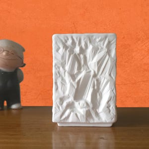 May include: A white rectangular vase with a textured, geometric design. The vase is on a wooden surface, with an orange background. A small figurine of a man in a black outfit is to the left of the vase.
