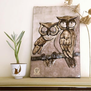 May include: A ceramic owl plaque featuring two owls perched on a branch, in shades of brown and cream. The plaque is rectangular and has a textured surface. A small potted plant in a white and brown pot is to the left.