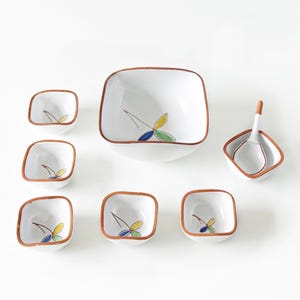 Puede incluir: Un juego de servir de cerámica blanca con diseño cuadrado. El juego incluye un cuenco grande, varios cuencos más pequeños y una cuchara. Cada pieza tiene un borde marrón y un diseño floral pintado con detalles amarillos, azules y verdes.