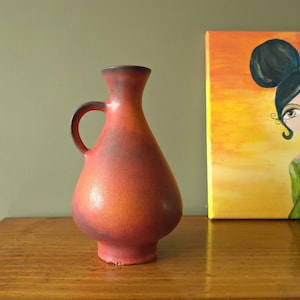 Puede incluir: Un jarrón de cerámica rojo y naranja con asa. El jarrón tiene un esmalte moteado y una forma redondeada.