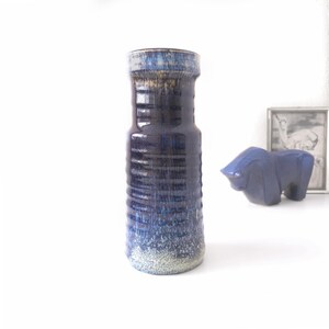 May include: A cylindrical ceramic vase featuring a deep navy blue glaze with horizontal ribbed textures. The top rim displays a lighter blue and cream drip effect, while the base transitions into a speckled cream and light blue finish.
