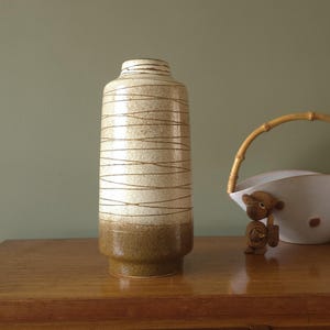 May include: A tall, cylindrical ceramic vase with a speckled off-white and brown color scheme. The vase features horizontal brown lines and a wider brown base. A decorative bowl with a bamboo handle and a small monkey figurine are also visible.