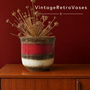 Op de afbeelding: Een keramische plantenpot met een horizontaal gestreept patroon in rood, donkerbruin en gebroken wit, gevuld met gedroogde bruine bloemstengels. De tekst VintageRetroVases staat in het wit boven de pot op een donkerrode achtergrond.
