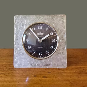 May include: A square, clear glass clock with a round clock face. The clock face is black with white numerals and hands. The clock reads "JUNGHANS electronic ATO-MAT". The clock is sitting on a wooden surface.