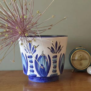 May include: A white ceramic planter featuring a hand-painted pattern of blue palm trees and stylized floral motifs. The vessel has a tapered shape with a solid blue base and a dark blue rim, containing a large, spiky purple allium flower head.
