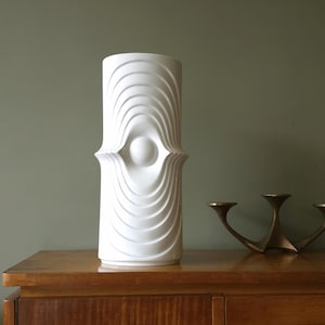 Könnte beinhalten: Weiße Keramikvase mit geometrischem Design. Die Vase hat eine zylindrische Form mit einem erhabenen, augenartigen Muster in der Mitte.