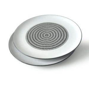 Op de afbeelding: Twee witte keramische borden met een zwart spiraalpatroon in het midden. De borden zijn op elkaar gestapeld, waarbij het bovenste bord iets schuin staat om het bord eronder te laten zien. Eenvoudig, modern ontwerp.