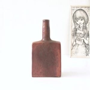 Puede incluir: Un jarrón de cerámica de forma cuadrada con un esmalte marrón y un acabado craquelado. El jarrón tiene un cuello estrecho y una base ancha.