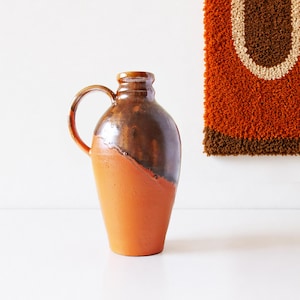 Könnte beinhalten: Ein Keramikkrug mit zweifarbigem Design, bestehend aus einer matten terrakotta-orangen Basis und einem glänzend dunkelbraun glasierten Oberteil. Das Gefäß hat einen bauchigen Körper, einen schmalen Hals und einen geschwungenen Henkel.
