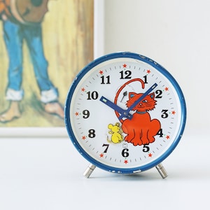 May include: A vintage, round alarm clock with a white face, blue hands, and a blue rim. The clock face features a cartoon cat and mouse, with red stars marking the hours. The clock has silver legs.
