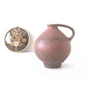 May include: A speckled, bulbous, ceramic pitcher with a handle, in shades of brown and pink. A circular framed floral arrangement with a gold rim is in the background. The pitcher has a wide base and a narrow neck.