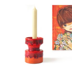 May include: A red and orange ceramic candle holder with a cream-colored candle. The candle holder has a unique hourglass shape. A painting with a girl holding flowers is in the background.
