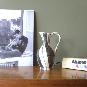 May include: A black and white striped ceramic pitcher with a handle. The pitcher is on a wooden surface, next to a black and white photograph of a man playing a trumpet and a book titled "Jazz & Cover".