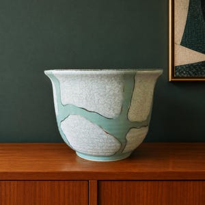 Op de afbeelding: Een keramische plantenpot met een brede, uitlopende rand en een afgeronde vorm. Het buitenoppervlak vertoont een opvallend patroon van witte, gebarsten textuurvlakken gescheiden door gladde, lichtgroenblauwe banden in een geometrisch ontwerp.