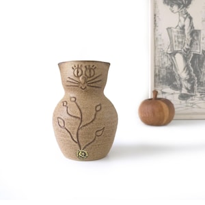 May include: A tan ceramic vase with a cat face design, featuring large eyes and whiskers. The vase has a textured surface and a brown plant design. A small wooden apple and a framed drawing are in the background.