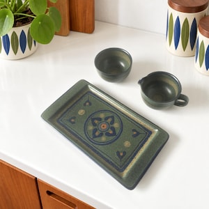 May include: A rectangular ceramic tray with a green, blue, and orange floral pattern, accompanied by a small matching bowl and a handled creamer. The set features a speckled finish and dark green glaze, coordinating with two cylindrical canisters.