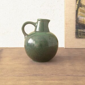 May include: A small, round ceramic pitcher with a glossy, mottled forest green glaze. The vessel features a curved handle on the left side and a short, wide spout at the top, showcasing a smooth, reflective surface with subtle variations in color.