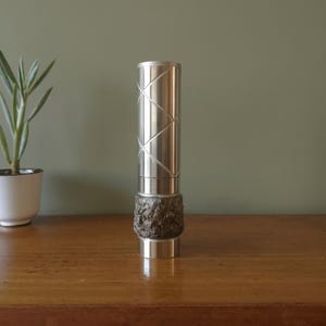 Può includere: Un vaso in metallo argentato con un motivo geometrico sulla parte superiore e una base testurizzata di colore grigio scuro.