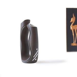 May include: A dark brown ceramic vase with a distinctive handle. The vase has a curved form, a white interior, and a white decorative pattern. The vase is approximately 20 cm tall and is set against a white backdrop.