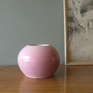 Peut inclure: Un petit vase rond en céramique rose avec une surface texturée. Le vase a une large ouverture et est posé sur une surface en bois. Une impression encadrée en noir et blanc est en arrière-plan.