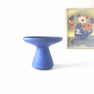 Puede incluir: Un cuenco de cerámica azul sobre pedestal con un borde circular ancho y una base cónica estrecha. La superficie del objeto presenta un acabado mate y texturizado en un color azul sólido y profundo, apoyado sobre una superficie plana.