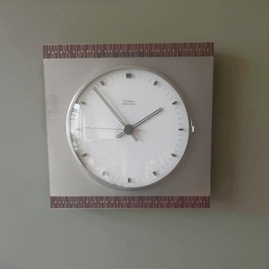 May include: A square wall clock with a white circular face, black hour and minute hands, and a thin red second hand. The clock features a silver-toned frame with dark brown patterned borders at the top and bottom. The text Diehl dielectron is printed in black on the centre of the white clock face.