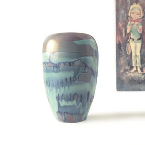 May include: A ceramic vase with a rounded silhouette featuring a metallic bronze base glaze overlaid with a vibrant turquoise blue drip pattern that cascades down the sides, creating a textured contrast between the smooth metallic and matte finish.