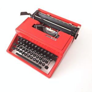 Peut inclure: Une machine à écrire Olivetti Dora rouge vintage avec des touches noires. La machine à écrire a un design classique avec un levier de retour de chariot et un mécanisme d'alimentation du papier. Les touches sont disposées en QWERTY. La machine est en bon état.