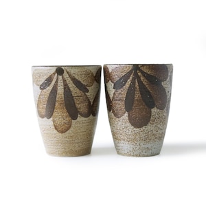 May include: Two ceramic tumblers with a beige and brown floral design. The tumblers have a textured surface and a cylindrical shape. The floral pattern is dark brown and covers a large portion of the tumbler's surface. The tumblers are suitable for beverages.