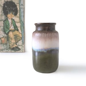 Puede incluir: Un jarrón de cerámica vintage con forma cilíndrica. La parte superior es de color marrón oscuro, que pasa a blanco y luego a una base verde oscuro. El jarrón tiene un acabado brillante y un borde ligeramente ensanchado. Una pintura de un niño está en el fondo.