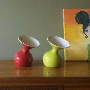 Puede incluir: Dos jarrones de cerámica. Uno es rojo, el otro verde lima. Ambos tienen una base bulbosa y un cuello curvo que conduce a una abertura blanca en ángulo. Los jarrones se exhiben sobre una superficie de madera.
