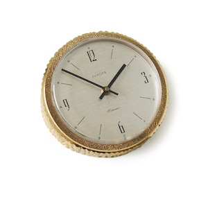 Puede incluir: Un reloj Europa dorado con un bisel texturizado y una esfera clara. El reloj presenta manecillas negras y números en las posiciones de las 12, 3, 6 y 9 en punto. La palabra "Europa" está impresa encima del centro.