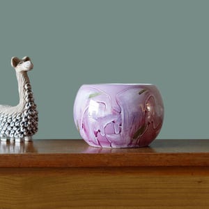 May include: A round, lavender-colored glass bowl with a swirling pattern of white, pink, and green. The bowl sits on a wooden surface, with a decorative llama figurine to the left. The bowl's design is abstract and artistic.
