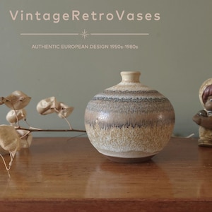 Può includere: Un vaso in ceramica rotondo con collo stretto, caratterizzato da fasce orizzontali di smalto beige, marrone e grigio scuro. Il testo "VintageRetroVases" e "AUTHENTIC EUROPEAN DESIGN 1950s-1980s" è visibile sopra il vaso nell'immagine.