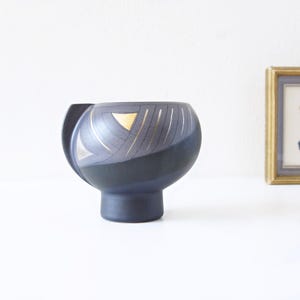 May include: A dark blue ceramic vase with a unique, rounded shape and a pedestal base. The vase features gold and silver geometric designs on the upper portion. The vase is displayed on a white surface, with a framed picture in the background.