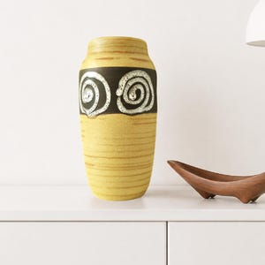 May include: A cylindrical ceramic vase with a yellow base featuring horizontal brown lines. A wide dark brown band encircles the upper section, decorated with two prominent white spiral patterns. The vase has a smooth, matte finish and a narrow opening.