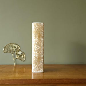 Op de afbeelding: Een hoge, rechthoekige witte vaas met een goudkleurig, webachtig patroon. De vaas staat op een houten oppervlak, met links een gouden metalen bladdecoratie. De achtergrond is een gedempt groen.