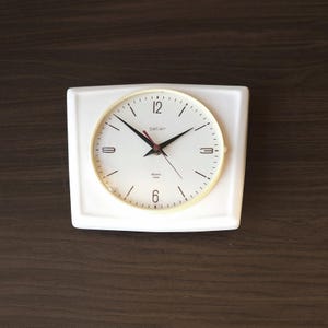 May include: A rectangular white wall clock with a circular face, featuring a cream-colored bezel, black hour and minute hands, a thin red second hand, and the text "peter" and "electric" printed in black on the white clock face dial.