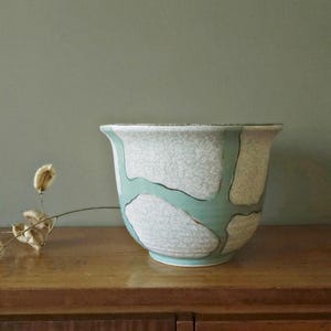 May include: A large, vintage ceramic planter with a wide, flared rim. The exterior features a textured, off-white background with a turquoise and black abstract design. The planter sits on a wooden surface.