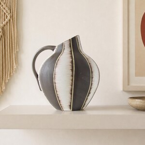 May include: A rounded ceramic pitcher featuring a matte dark charcoal finish with vertical white stripes that have a feathered, brushstroke texture. The vessel has a curved handle on the left side and an asymmetrical, slanted top opening.