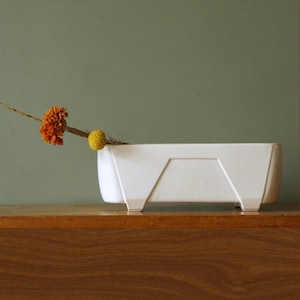 Può includere: Una fioriera rettangolare bianca in ceramica con un design geometrico. La fioriera contiene un fiore arancione secco e un fiore giallo a forma di palla. La fioriera è posizionata su una superficie di legno su sfondo verde.