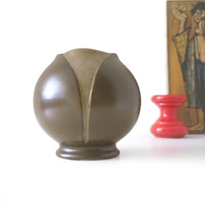 Könnte beinhalten: Eine braune Keramikvase mit einer einzigartigen, abgerundeten Form und einem vertikalen, helleren Akzent. Die Vase hat einen ausgestellten Sockel und eine geteilte Oberseite. Ein kleines, rotes Dekorationsobjekt befindet sich im Hintergrund.