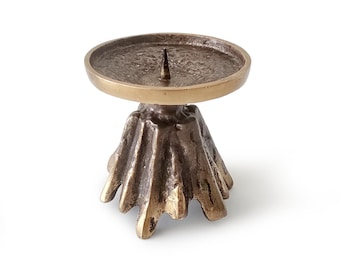 May include: A bronze-toned metal candle holder featuring a circular plate with a central spike for securing a candle. The base has a textured, organic, and flared design with multiple vertical ridges that extend outward toward the bottom edge.