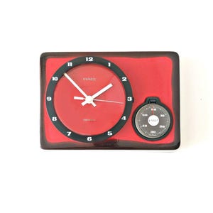 Puede incluir: Un reloj Kienzle vintage rojo y negro con cronómetro. La esfera del reloj es roja con números y manecillas negras. El cronómetro es blanco y negro. El reloj tiene la palabra "KIENZLE" en la esfera.