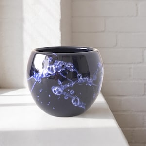 Op de afbeelding: Een ronde keramische plantenpot met een glanzende donker marineblauwe afwerking. Het oppervlak is voorzien van een opvallend kristallijn glazuurpatroon in tinten helder violet en wit, wat een organisch, gemarmerd effect geeft aan de buitenkant.